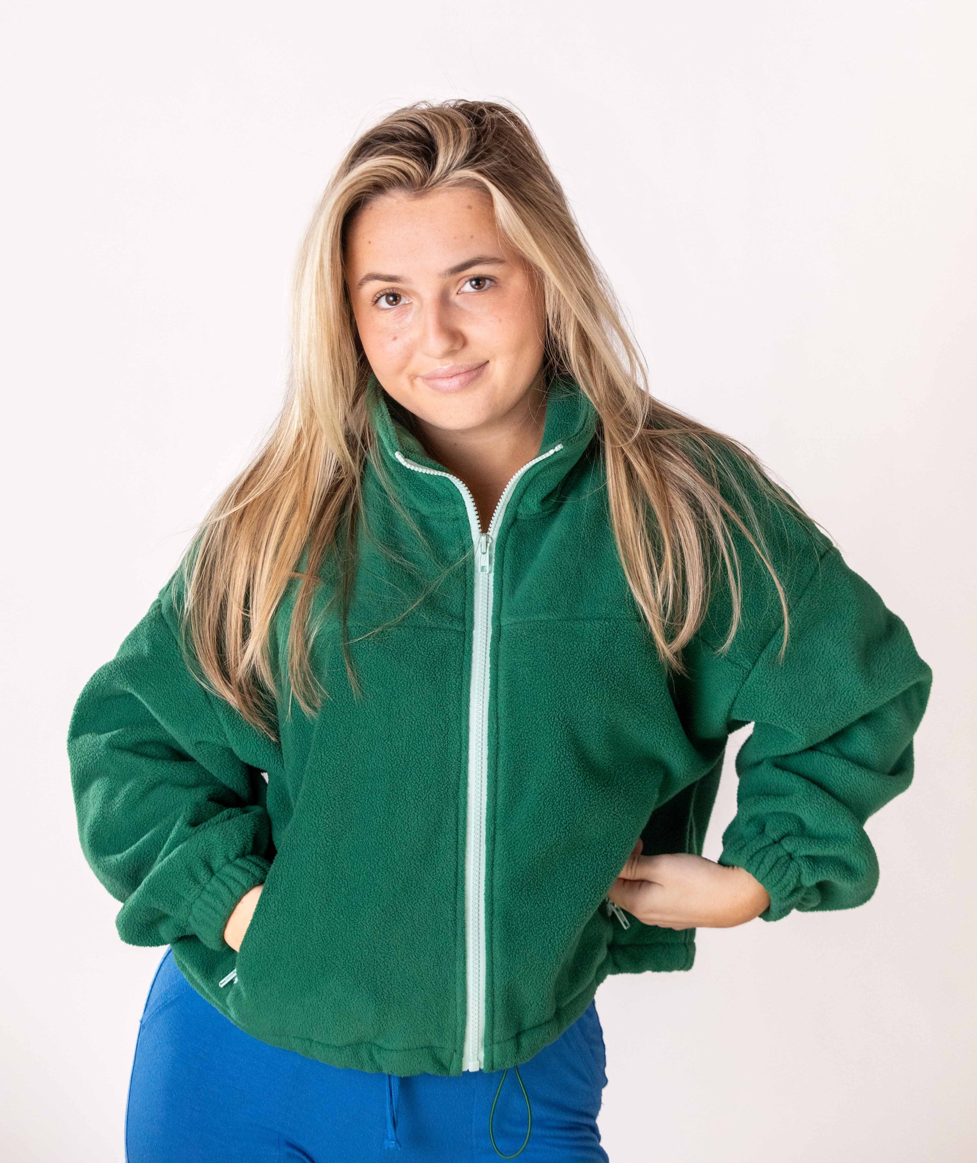 Woman wearing a green pullover fleece jacket with a mint color zipper on a plain background. The jacket has pockets with same mint color detail with pocket zipper. The jacket has a collar and pull string fastener at button. 