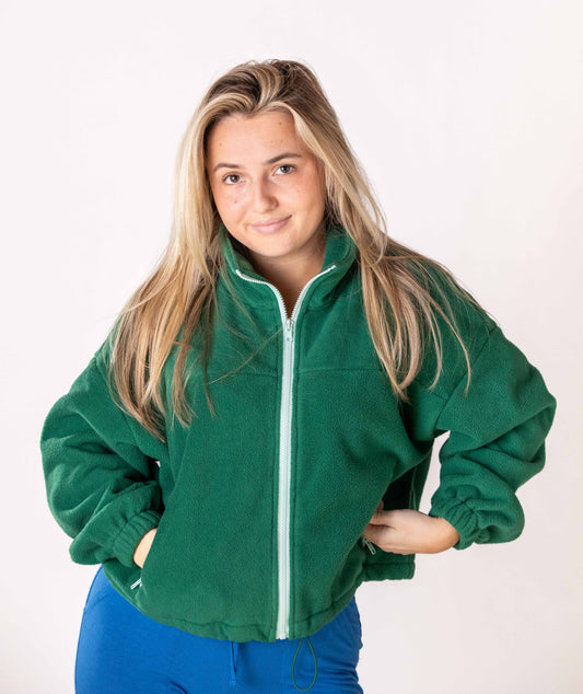 Woman wearing a green pullover fleece jacket with a mint color zipper on a plain background. The jacket has pockets with same mint color detail with pocket zipper. The jacket has a collar and pull string fastener at button. 