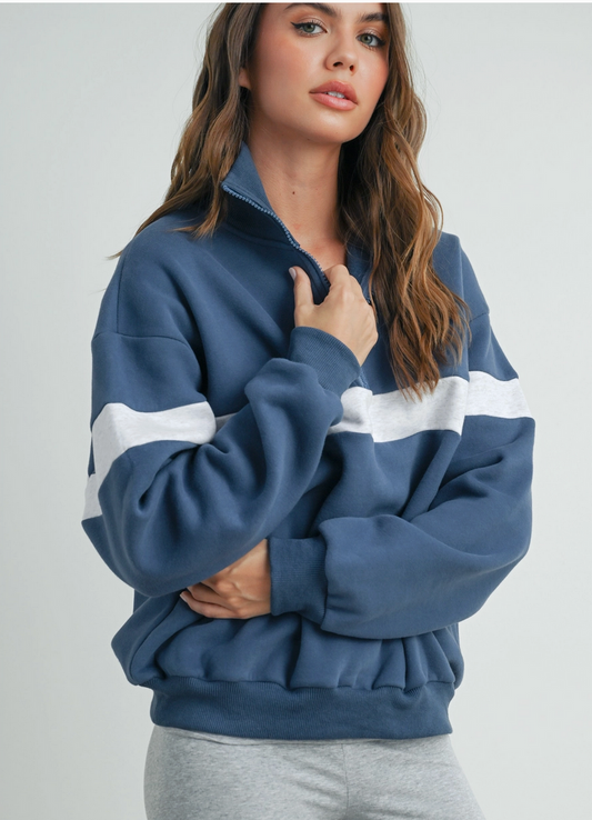 Woman wearing a blue and white sweatshirt on a light gray background. the sweatshirt is light navy blue with gray/white stripe in center. Half zip with high collar. 